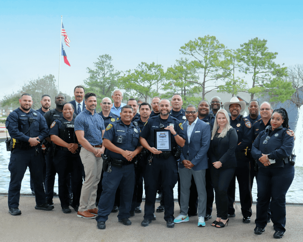 Group of police officers, sheriff deputies and professionals with the National Night Out award from 2022. Ourdoor shot with blue sky, green trees and US and Texas flags flying. The group is standing in front of a water feature. 