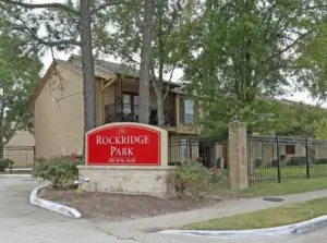 Photo of entrance to Rockridge Park apartments with bright red entrance sign and tan apartment building with beautiful trees and landscaping. 