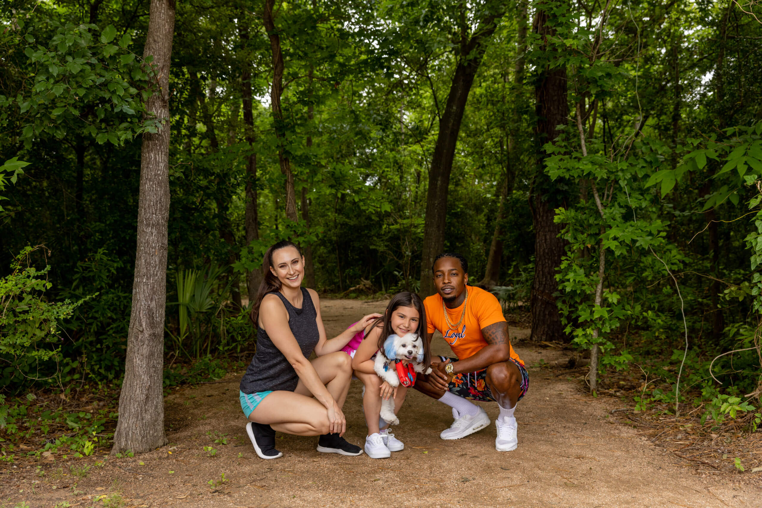 Family at the park! Mom is on the left kneeling down, daughter is in the middle holding a cute small dog and dad is on the right smiling on a manicured trail with woods in the background.