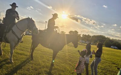 Two police officers on horseback are silhouetted against the setting sun, interacting with a small group of children in an open grassy field. The sunlight creates long shadows, and the group seems engaged in conversation as they enjoy the evening event.