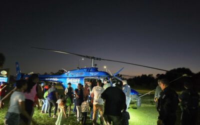 A crowd of people, including children and families, gather around a blue sheriff helicopter on display at night. The helicopter is illuminated by the surrounding lights, and people wait to get a closer look, with some taking photos.