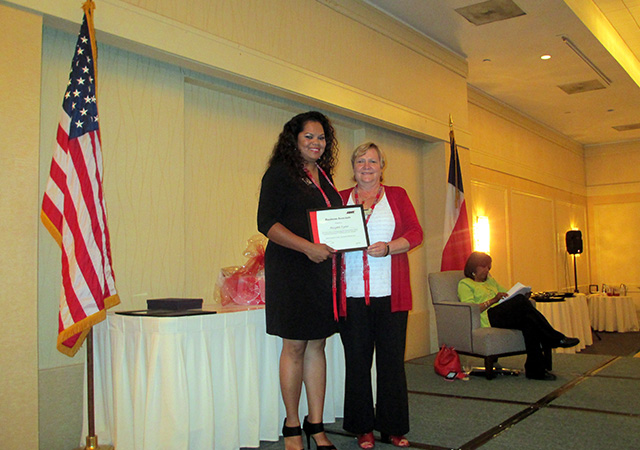One woman presents a certificate to the other at an award ceremony, with American and Texas flags visible behind them.