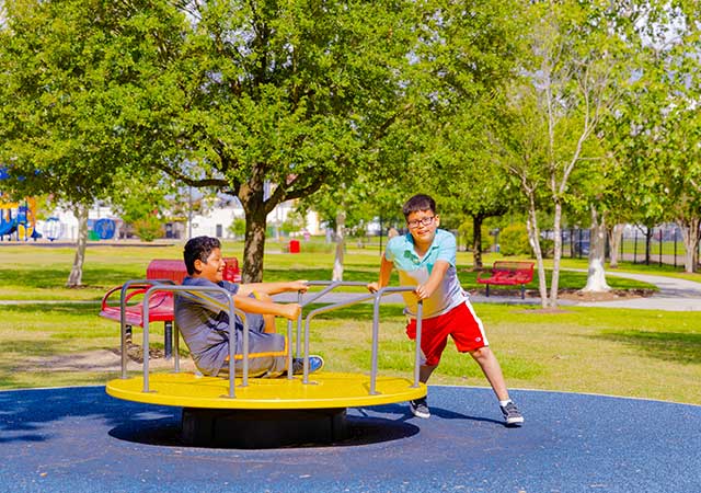 Two children play on a round yellow playground spinner at Buckboard Park, with one child pushing while the other rides, surrounded by rubberized surfacing and trees.