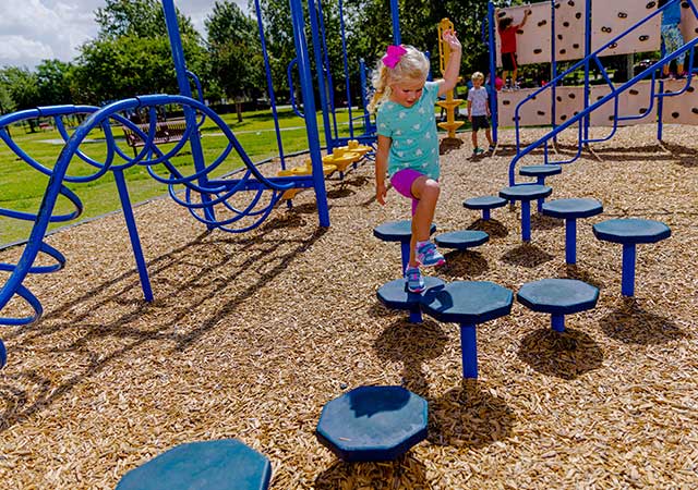 A young child steps across round balance pods on a playground at Buckboard Park, with climbing equipment and wood-chip surfacing visible around the play area.