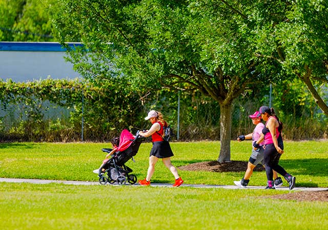 Adults walk along a paved trail at Buckboard Park, including one person pushing a stroller, as trees provide shade along the grassy park setting.