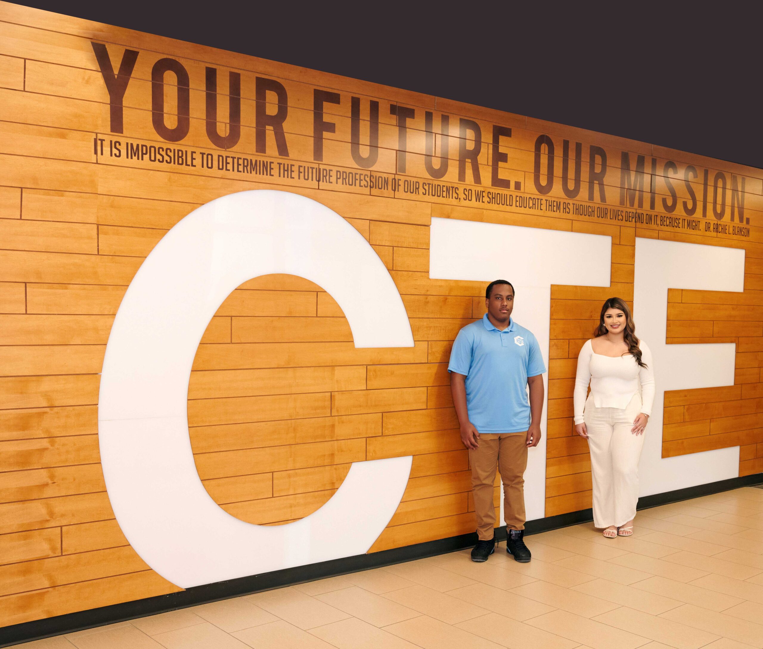 Two students stand side by side in front of a large wood-paneled wall that says CTE – Your Future. Our Mission.
