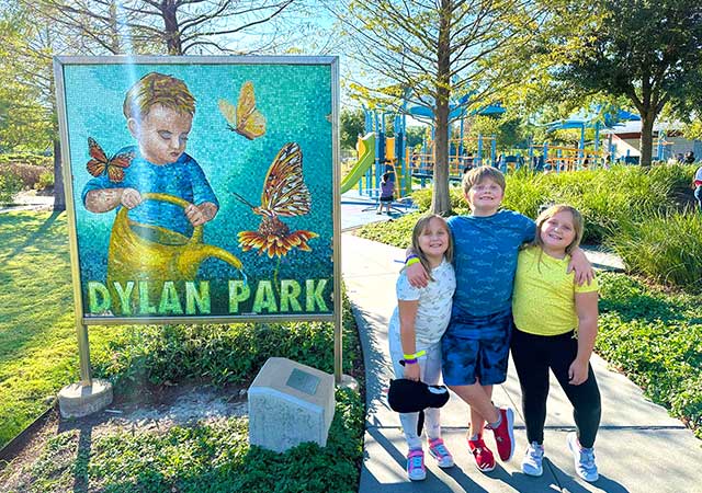Three children stand smiling beside a mosaic sign reading Dylan Park, with playground equipment, trees, and landscaped greenery visible along a paved park walkway.