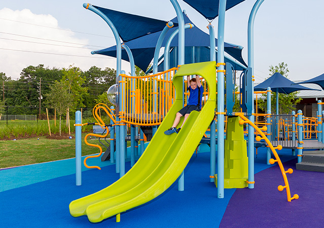 Child slides down a bright green playground slide at Dylan Park, featuring colorful play structures, climbing elements, and overhead shade canopies.