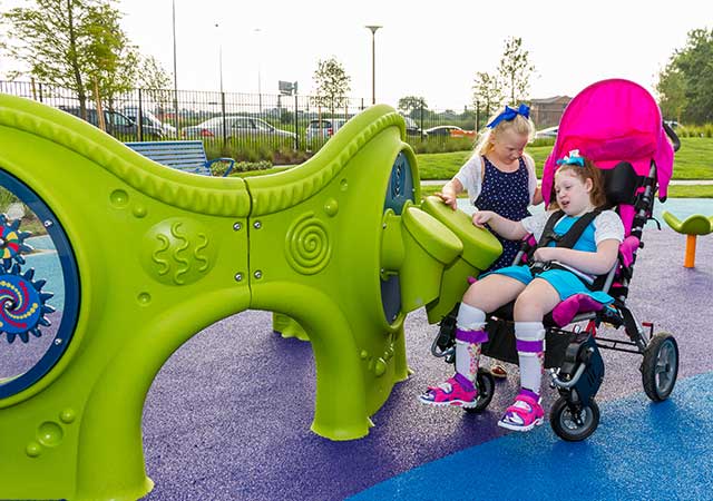 Two children play together at an inclusive playground feature at Dylan Park, with one child seated in a stroller-style mobility chair beside bright green adaptive equipment.