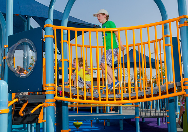 Children play on colorful playground equipment at Dylan Park, featuring elevated walkways, slides, climbing structures, and shade canopies.