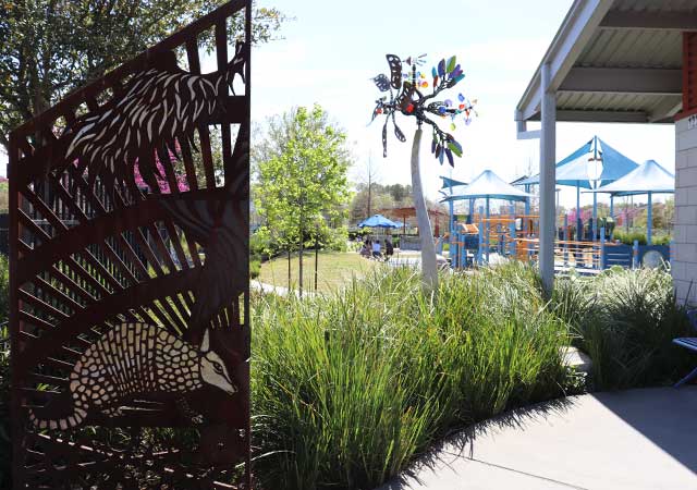 A decorative metal art panel with nature-inspired cutout designs stands along a landscaped walkway at Dylan Park, with playground structures, ornamental grasses, and shade structures visible nearby.