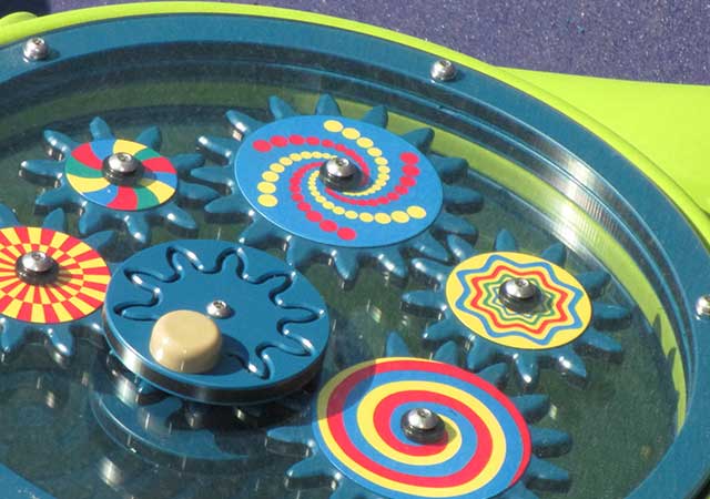 A close-up of a colorful interactive playground gear panel at Dylan Park shows multiple spinning gears with bright patterns designed for sensory play.