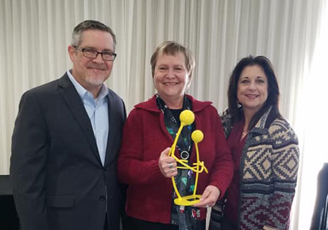 A woman in the center holds a bright yellow sculptural award, flanked by a man and another woman, all smiling indoors.
