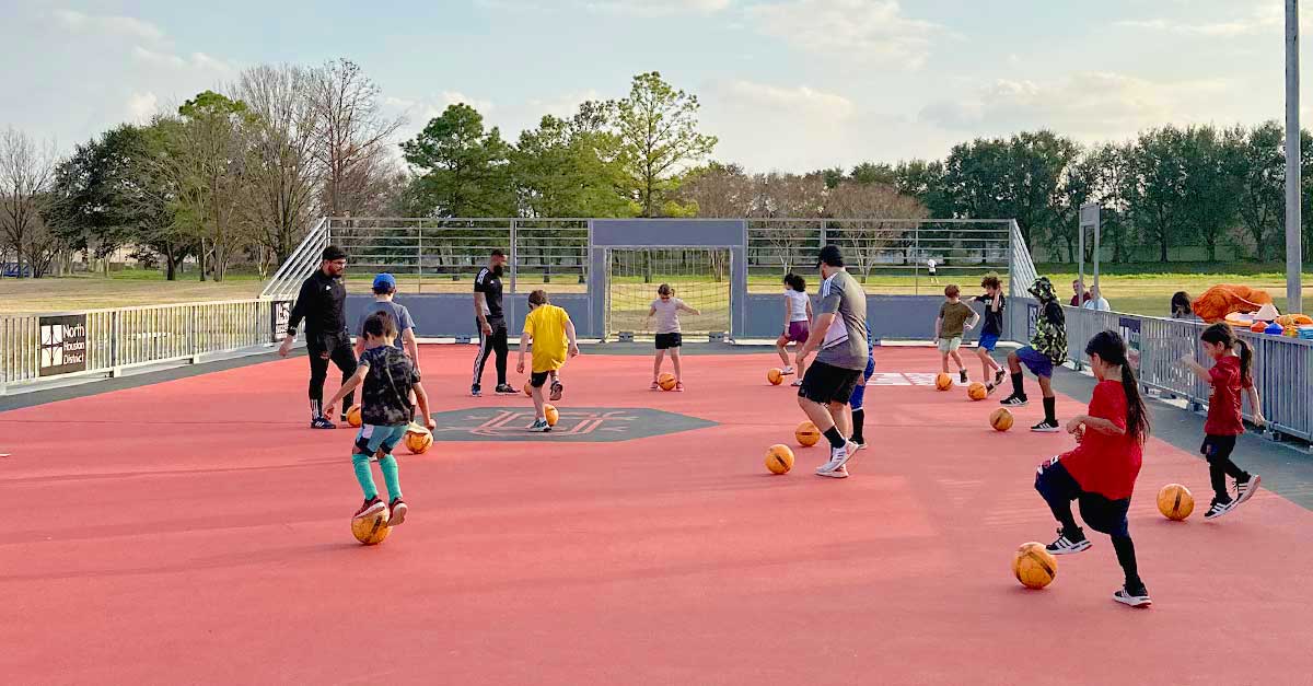 Children practice soccer drills with orange balls on a red mini pitch at Wussow Park, guided by coaches during a youth clinic hosted by the Houston Dynamo