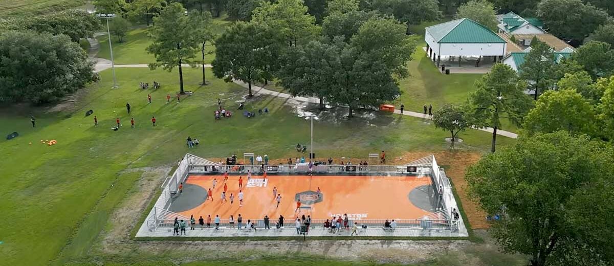 Aerial view of a youth soccer clinic at the mini pitch in Wussow Park, with players on the court, spectators around the field, and park pavilions in the background.