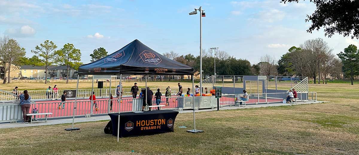 A Houston Dynamo FC tent and table are set up near a red mini soccer pitch at Wussow Park, where children and families gather for a youth soccer clinic.