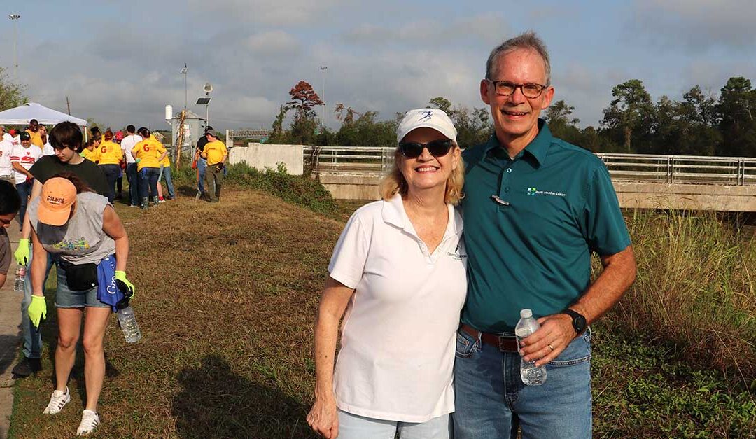 Two people are smiling and posing outdoors at a community event. The woman, wearing sunglasses, a white polo shirt, and a white cap, stands next to a man in a teal polo shirt with the North Houston District logo. He is holding a water bottle. Behind them, a group of volunteers in brightly colored shirts is gathered near a grassy area and a bridge under a partly cloudy sky.
