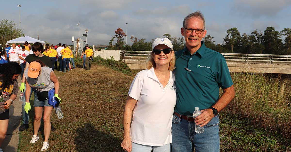 Two people are smiling and posing outdoors at a community event. The woman, wearing sunglasses, a white polo shirt, and a white cap, stands next to a man in a teal polo shirt with the North Houston District logo. He is holding a water bottle. Behind them, a group of volunteers in brightly colored shirts is gathered near a grassy area and a bridge under a partly cloudy sky.
