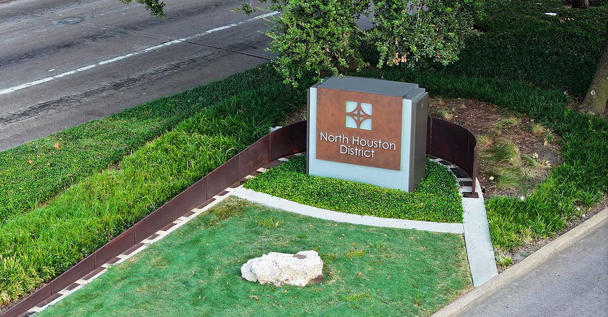Gateway monument sign for the North Houston District along a landscaped median with lush green grass and shrubs. The modern brown-and-gray structure displays the District logo prominently.