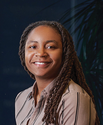 Hershiira Rogers Boone Headshot Headshot of Hershiira Boone-Rodgers smiling, wearing a striped blouse, in front of a dark blue background with a leafy plant in soft focus.