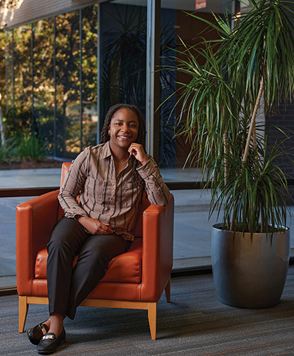 Hershiira Rogers Boone Headshot2 Hershiira Boone-Rodgers sits comfortably in an orange armchair, smiling, with one hand resting on her chin. She wears black pants and a beige button-up shirt with black stripes. A large potted plant stands nearby.