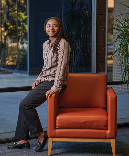 Hershiira Rogers Boone Headshot3 Hershiira Boone-Rodgers, wearing black pants and a beige button-up shirt with black stripes, sits on the arm of a burnt-orange chair near a window with greenery outside.