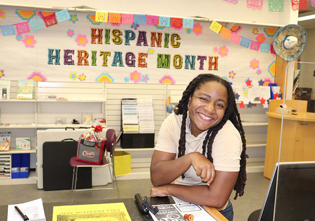 Hershiira Rogers Boone Library Desk Hershiira Boone-Rodgers smiles while leaning on a counter inside Aldine Library, with a Hispanic Heritage Month banner and colorful decorations behind her.