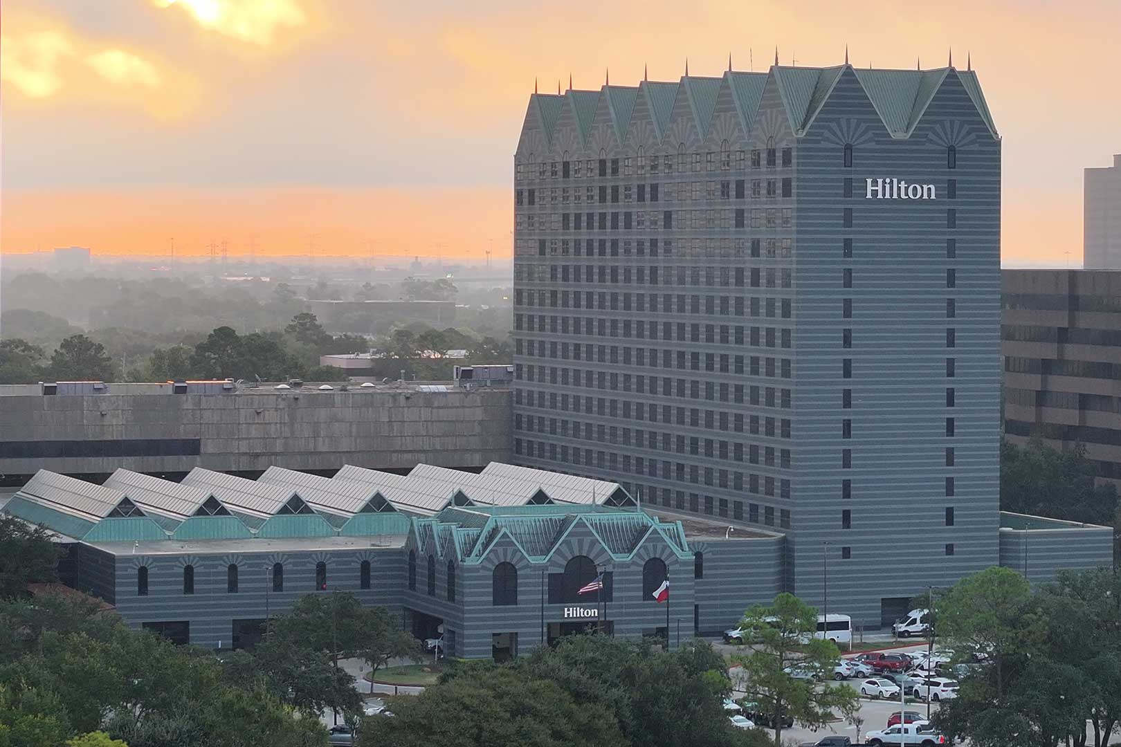 A wide view of the Hilton Houston North hotel surrounded by office buildings, with a dramatic sunrise casting light through the clouds in the background.