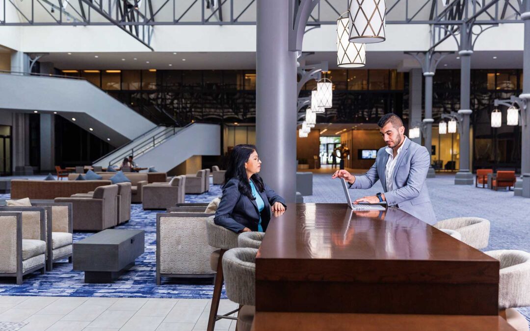 Two professionally dressed people talk commercial real estate at a tall table in a spacious, modern hotel lobby with stylish lighting, blue carpet, and contemporary lounge seating in the background.