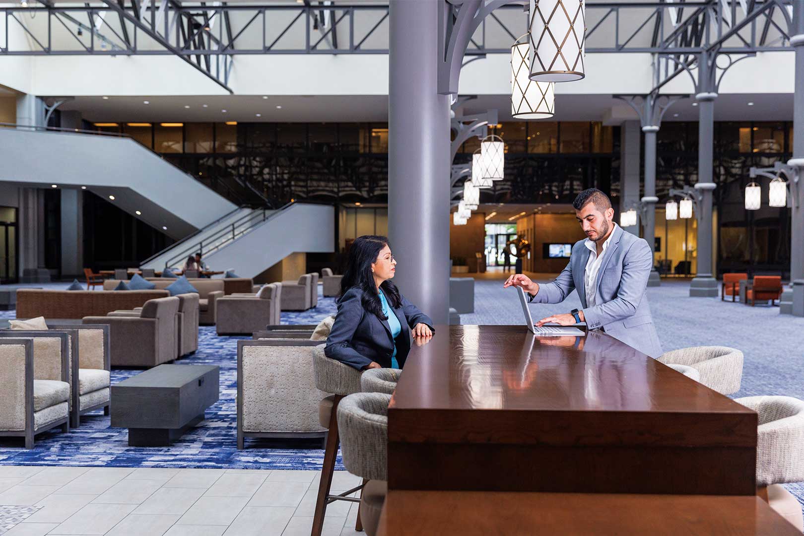 Two professionally dressed people talk commercial real estate at a tall table in a spacious, modern hotel lobby with stylish lighting, blue carpet, and contemporary lounge seating in the background.