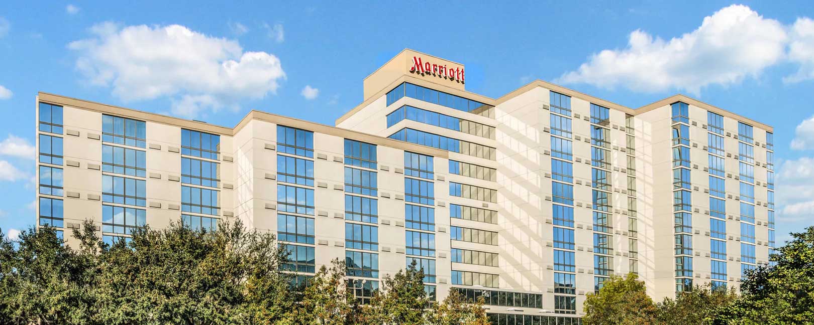 A bright daytime photo of the Houston Marriott North hotel featuring its modern glass windows, cream-colored exterior, and blue sky with scattered clouds overhead.