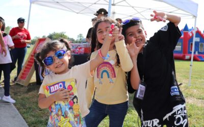 Three children smile and hold up small items while posing for a picture at the event. They stand in front of a tent, with one girl wearing sunglasses and holding a coloring book. A bounce house can be seen in the background, and the sunny day adds to the cheerful atmosphere.