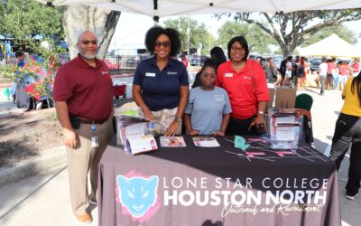 Four individuals stand behind a table under a tent, representing Lone Star College Houston North. They smile while standing next to brochures and promotional materials, with a large tablecloth displaying the school’s logo and outreach information.