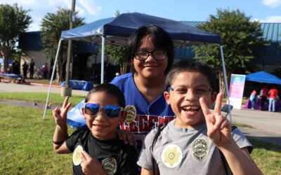 Three young boys pose for the camera, flashing peace signs and wearing sticker badges on their shirts. One boy wears sunglasses, and all three are smiling. The background shows event tents and activities under a sunny sky.