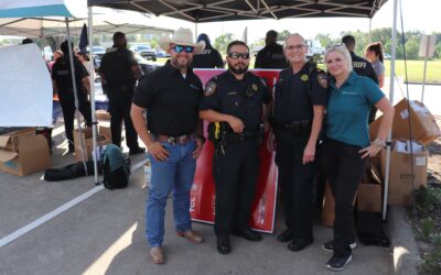 Four individuals, including two police officers, a man in a cowboy hat, and a woman in a polo shirt, pose together under a tent. They smile for the camera with event activities and other participants visible in the background.