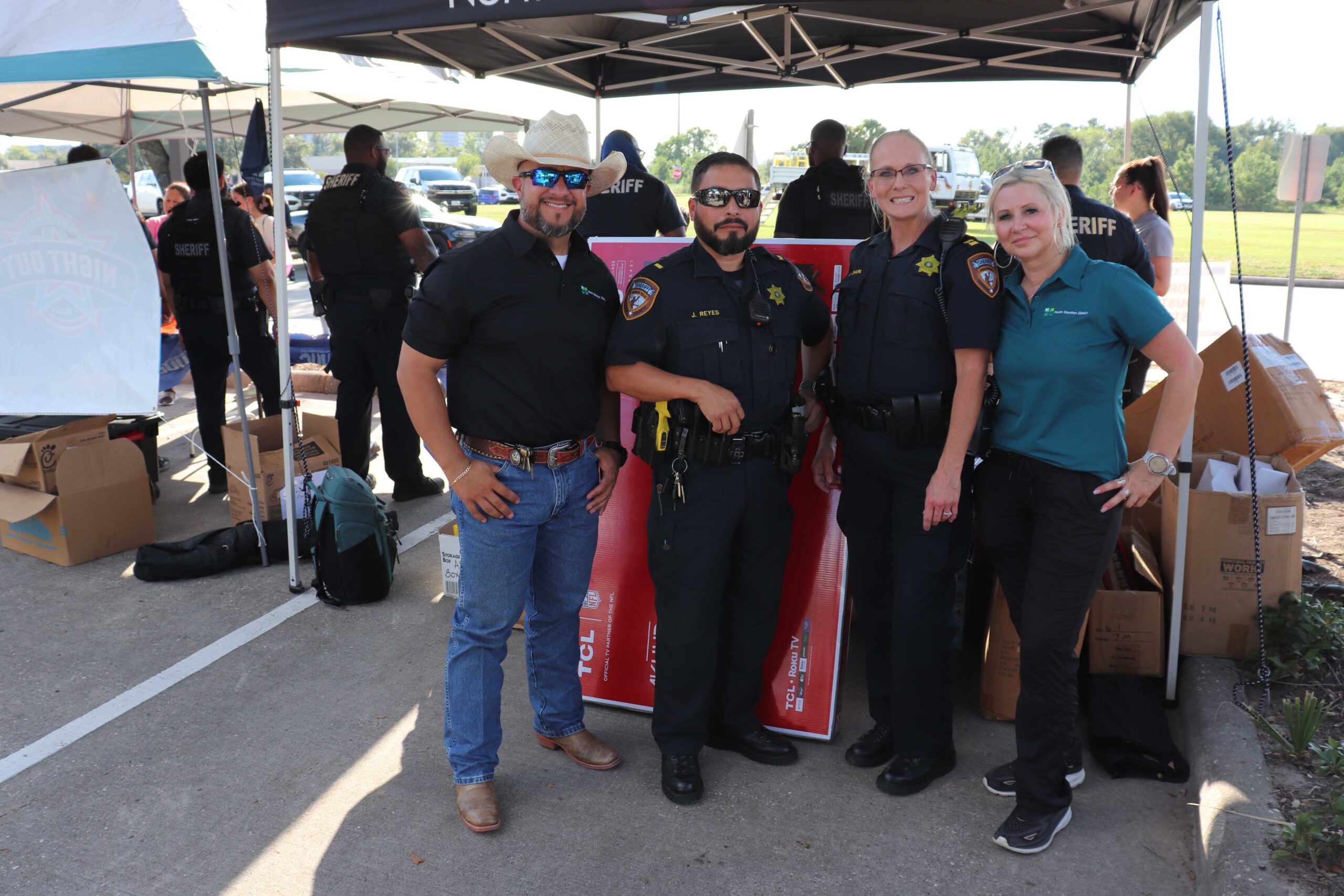 Four individuals, including two police officers, a man in a cowboy hat, and a woman in a polo shirt, pose together under a tent. They smile for the camera with event activities and other participants visible in the background.