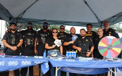 A group of eight uniformed officers from the Sheriff's Department pose together at a National Night Out booth. The booth is decorated with a blue tablecloth displaying the event's logo and promotional items, including sunglasses, water bottles, and a colorful spinning wheel. The officers are smiling, standing together under a tent.