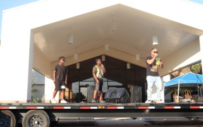 Three young performers stand on an outdoor stage in front of a building with a tall white facade. One performer is holding a microphone and speaking while the other two stand on either side. They are dressed casually, and the stage is set up for entertainment at the National Night Out event.
