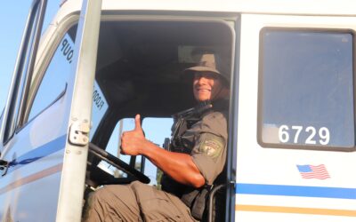 : A law enforcement officer in tactical gear, sitting inside a large vehicle and giving a thumbs-up. The officer is smiling, and the door of the vehicle has the number "6729" and an American flag decal on it.