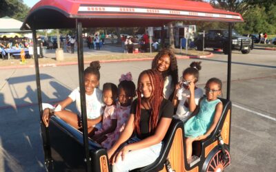 A group of seven young girls, all smiling, are seated inside a small, colorful train ride cart. The train is stationary on a paved area during an outdoor event. Behind them are various event booths, and the warm sunlight adds a golden glow to the scene. Some of the girls are wearing glasses, and everyone appears to be enjoying the ride.