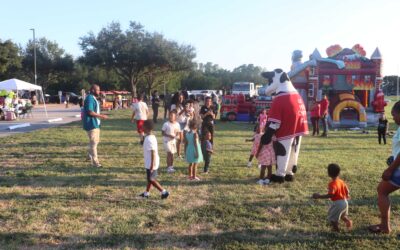 A large cow mascot, dressed in a red shirt that says "Eat Mor Chikin" from Chick-fil-A, stands in a grassy field with children gathered around. The cow is waving and interacting with the community, and a young girl in a pink dress with heart patterns is standing next to the mascot, looking at the camera. In the background, people are enjoying the event, and there are inflatable structures and booths scattered around the field.