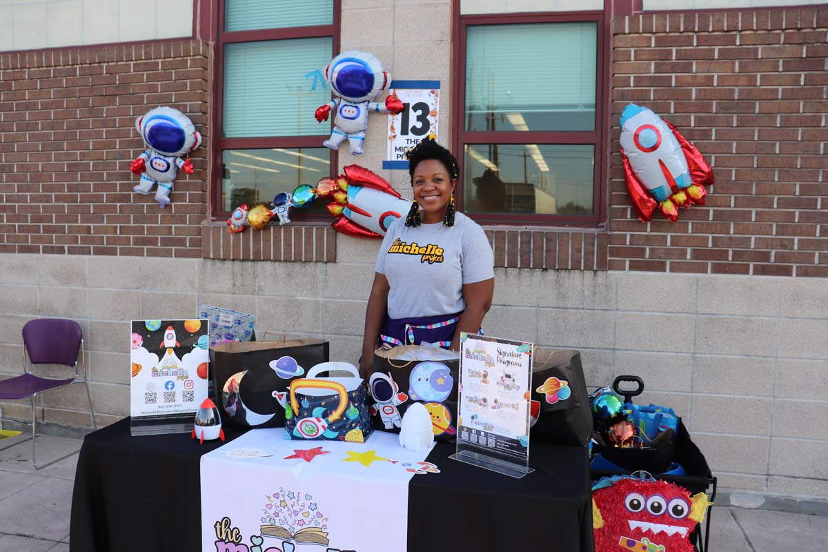 A woman stands behind a space-themed booth for "The Michelle Project" with space decorations, including astronaut balloons and rockets. The table displays various colorful items related to outer space.