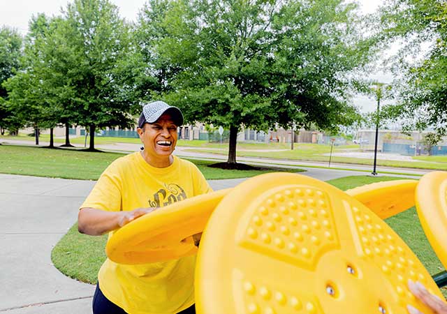 A person smiles while using yellow outdoor fitness equipment at Ida Gaye Gardens, with paved walkways, trees, and additional exercise stations visible in the park setting.