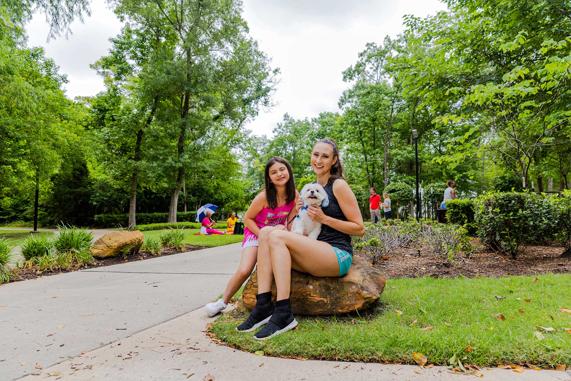 A smiling woman and child sit on a rock with a small white dog, surrounded by trees and people enjoying the park.