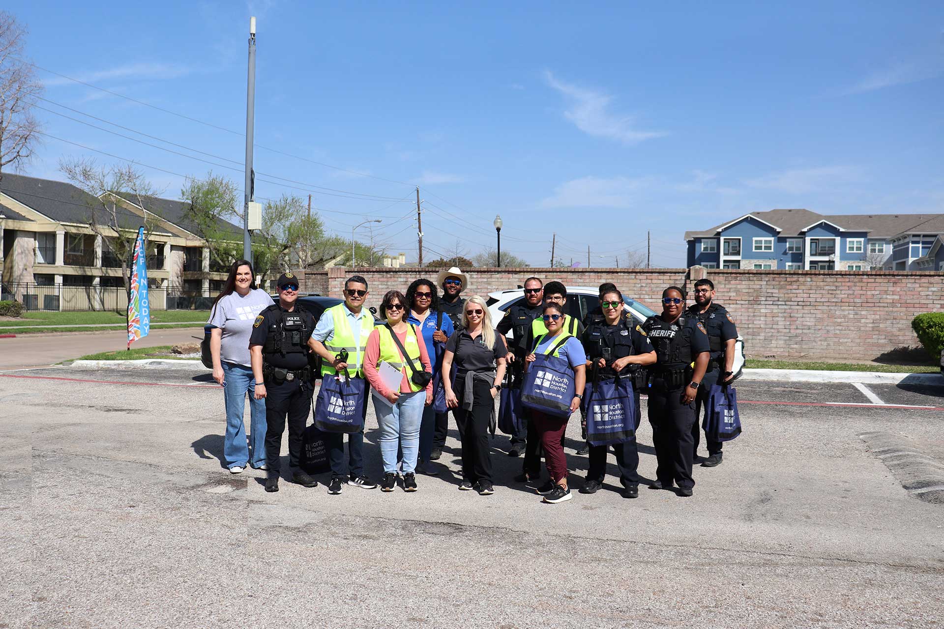 Large group photo of law enforcement and volunteers holding blue North Houston District bags in an apartment parking lot.