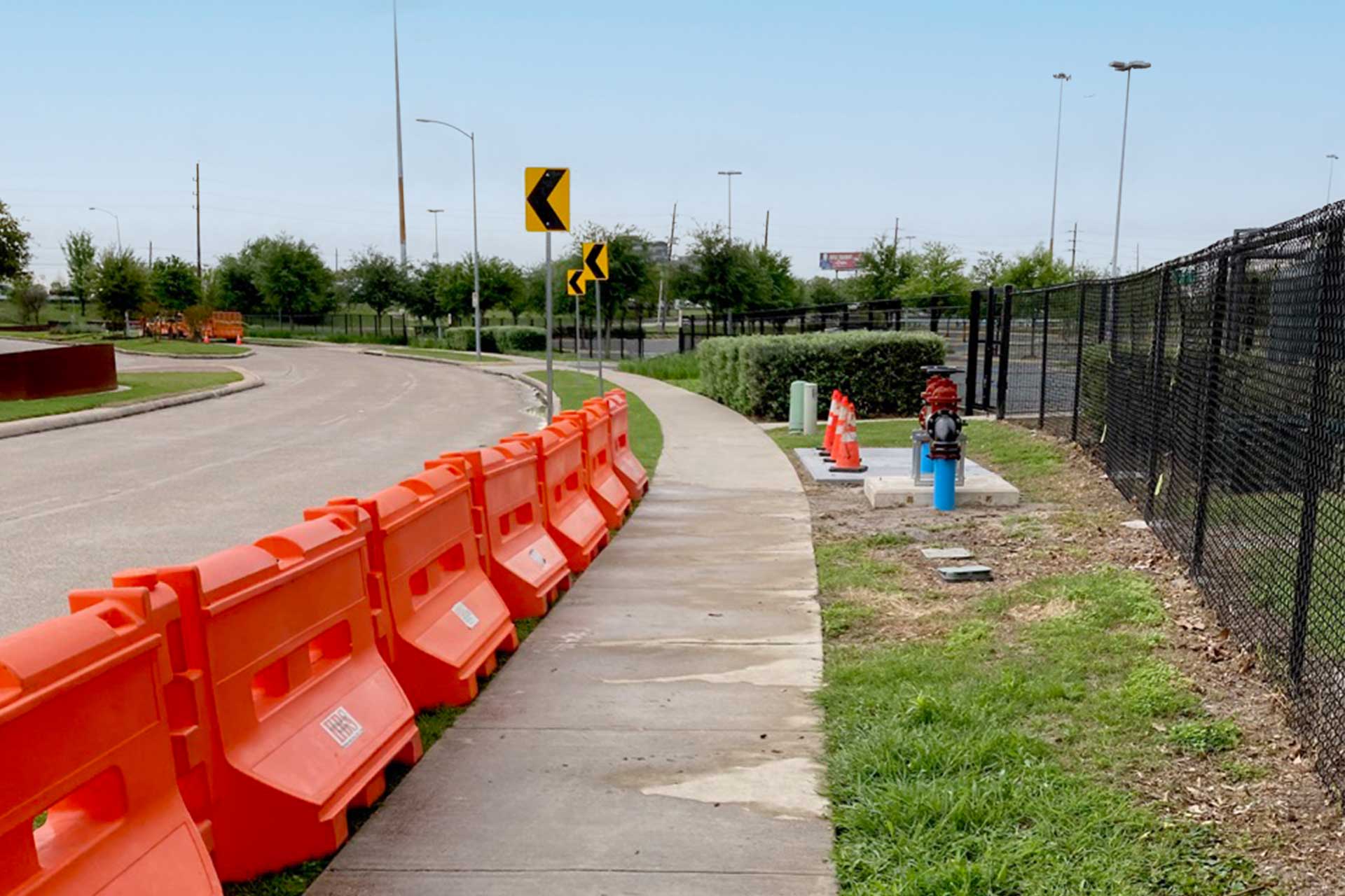 A curved sidewalk runs alongside a road with orange construction barriers lined up on its left side. On the right, a black chain-link fence borders a grassy area. A fire hydrant and utility equipment are visible next to the sidewalk. Traffic warning signs can be seen in the background near the curve in the road. The sky is clear, and the area appears to be under construction or improvement.