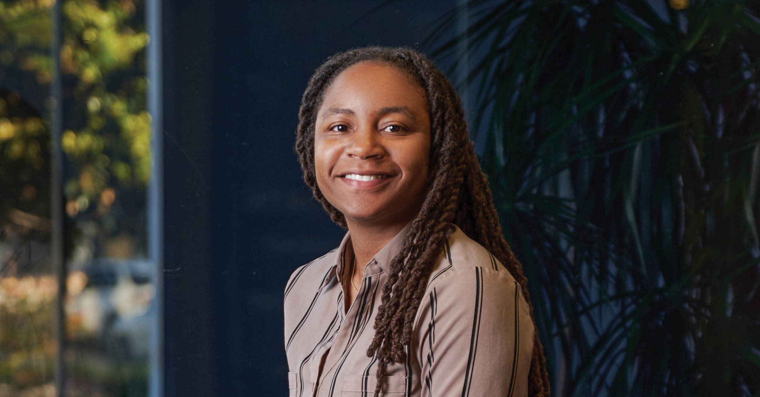 Close-up of Hershiira Boone-Rodgers smiling, wearing a beige button-up shirt with black stripes, with blurred greenery in the background.
