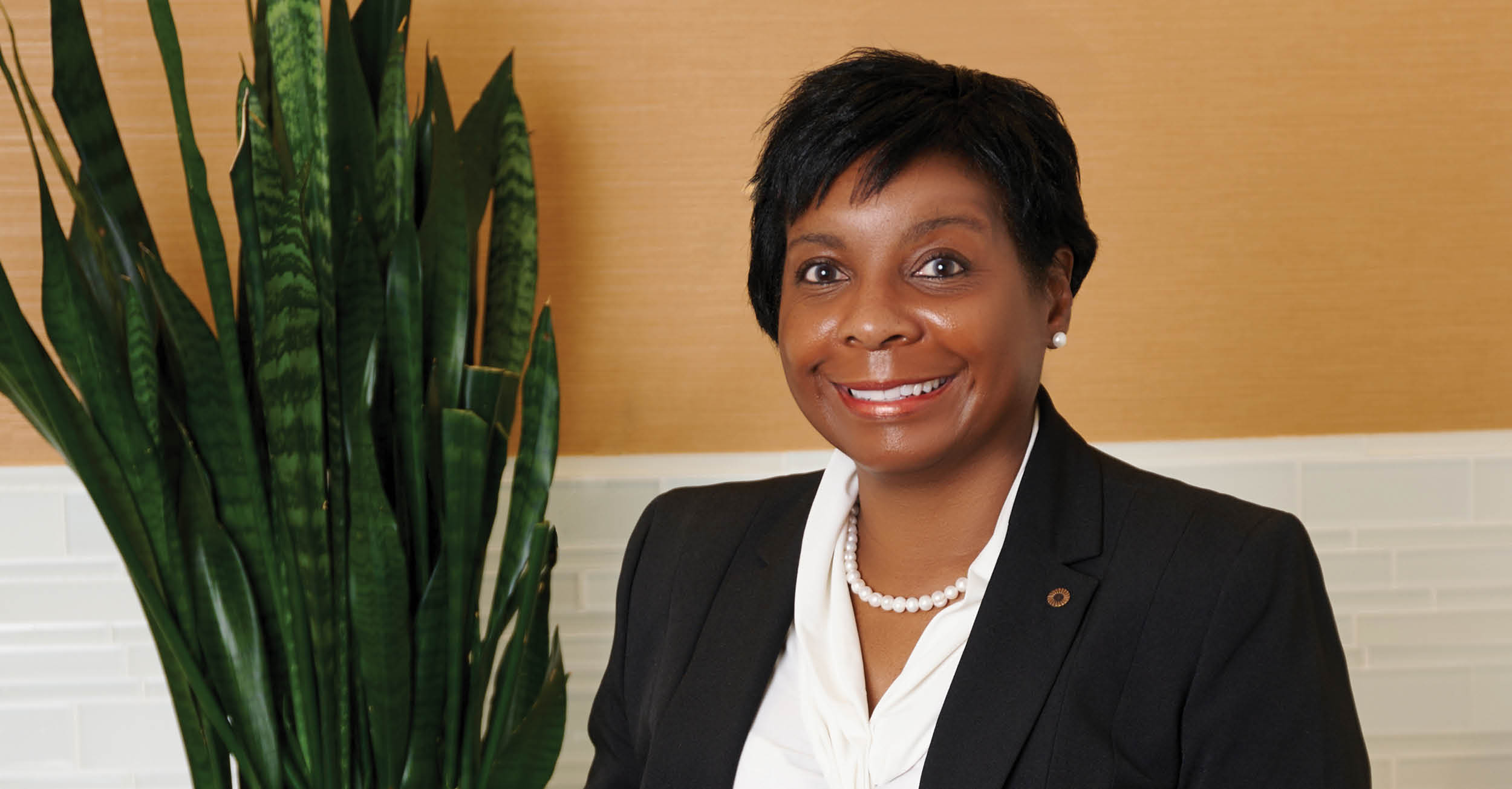 Tina Poindexter smiles in a business portrait, wearing a dark blazer and pearl necklace, standing beside a tall green plant against a warm-toned wall.