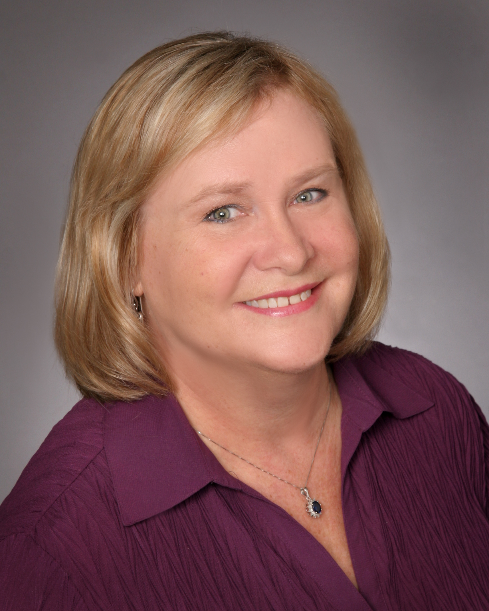 Professional headshot of Margaret Eyster in a plum blouse with a pendant necklace, smiling in front of a gray background.