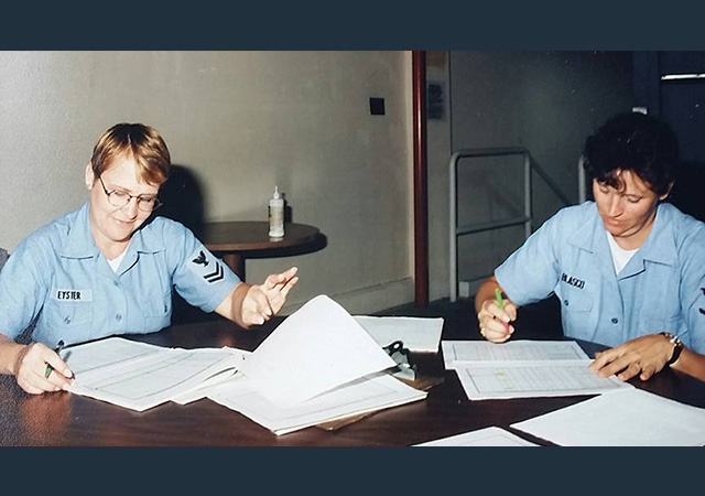 Two women in Navy uniforms work at a table covered with papers and folders, reviewing documents.
